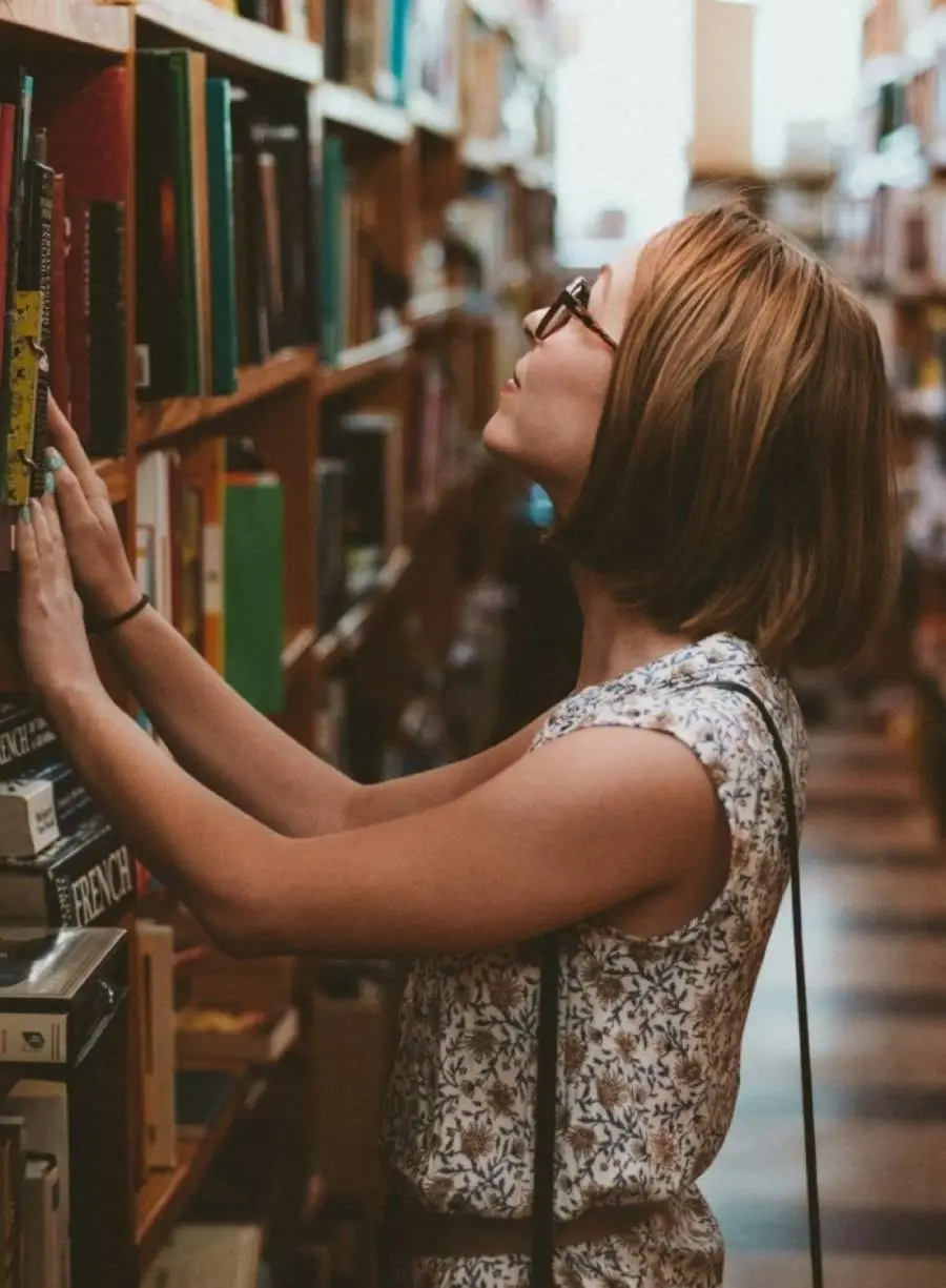 A woman wearing glasses browsing through a hallway of books in a library.