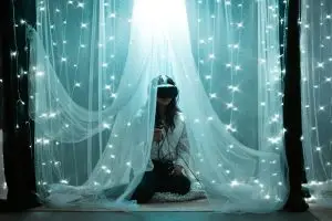 A woman in VR goggles draped with white curtain with white fairy lights.