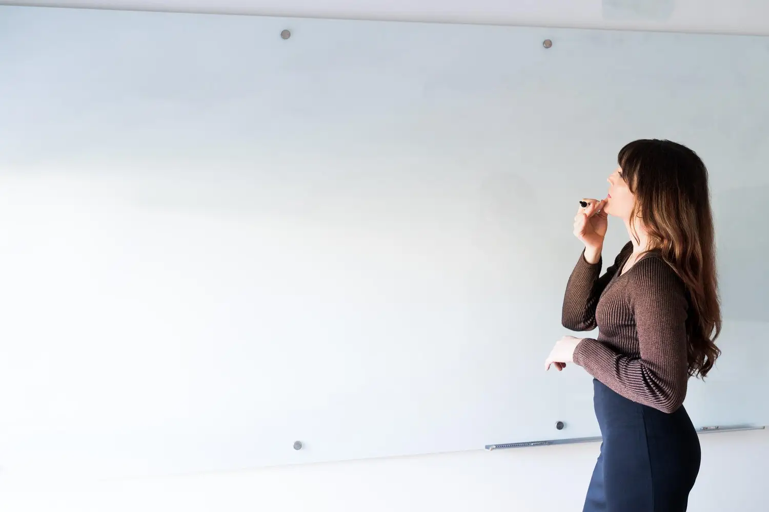 A woman thinking with her hand on her chin while facing a whiteboard.