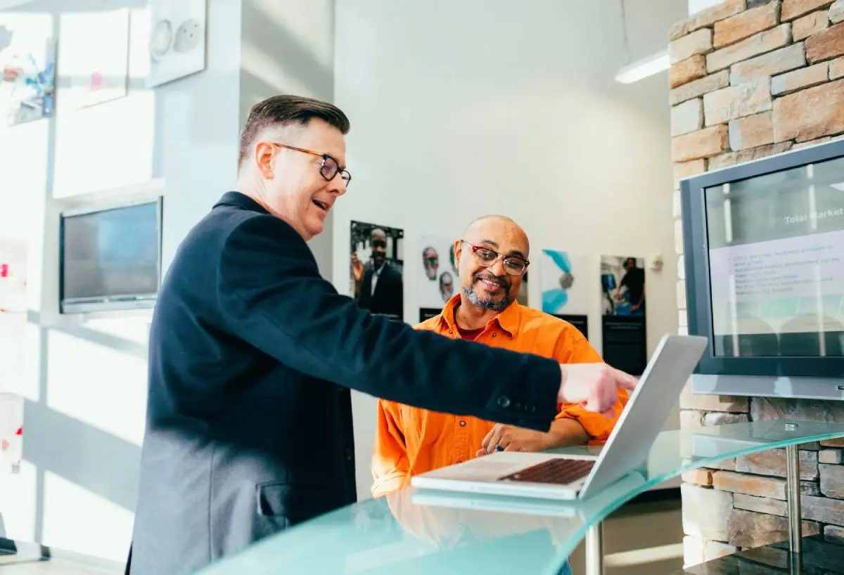 A man in a suit is pointing at a screen while the man in an orange clothing is smiling with the looks of content.