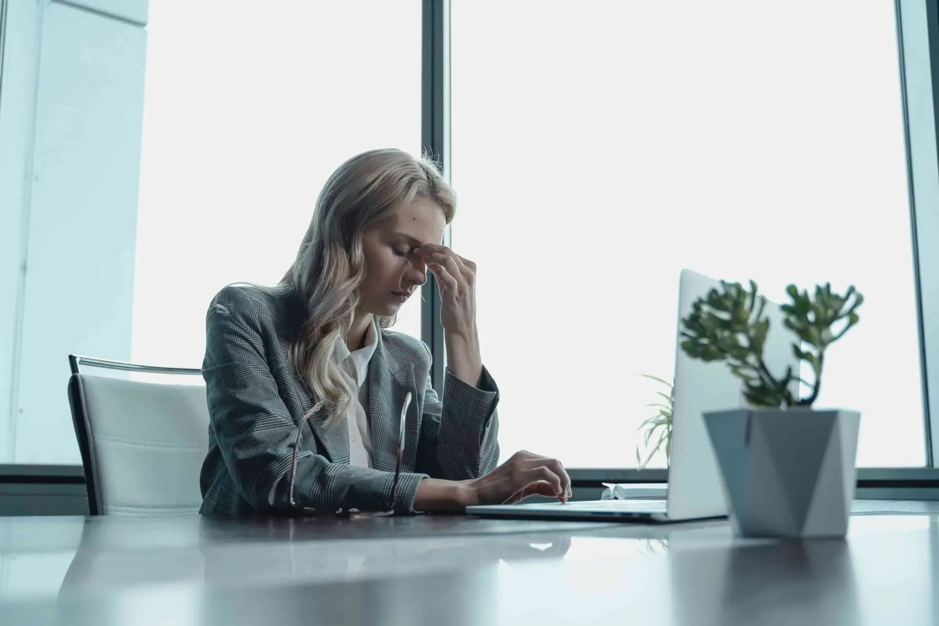 A woman sitting in an office facing her laptop while pressing the center of her forehead because of stress.